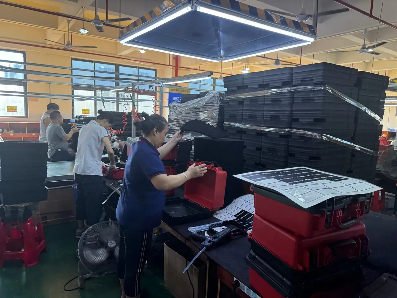 Artillery Shell Box: a worker in a blue shirt over a red case, printed label sheets on the bench, wrapped black cases on pallets behind