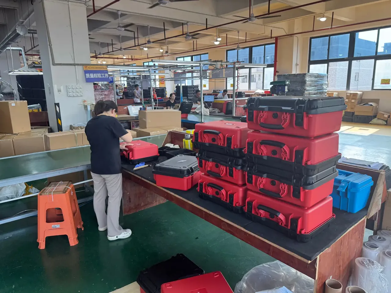 Case Assembly: stacks of red and black plastic tool cases on a wooden bench, black overmoulded corner caps on each lid, a worker in a dark top closing a case beside a conveyor