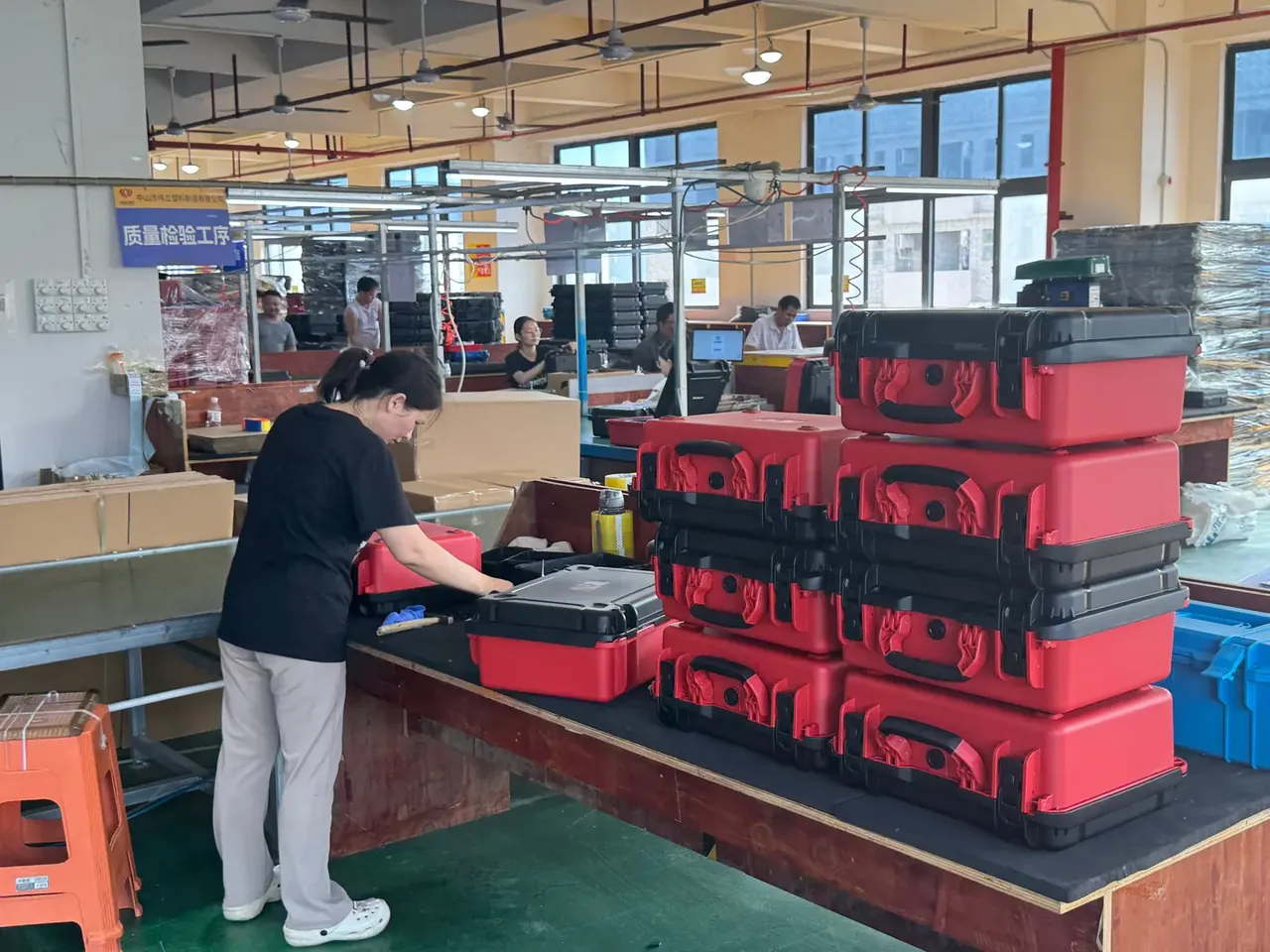 Case Assembly bench: a worker in black lifting the lids of a red and a grey case, stacks of red cases with black latches