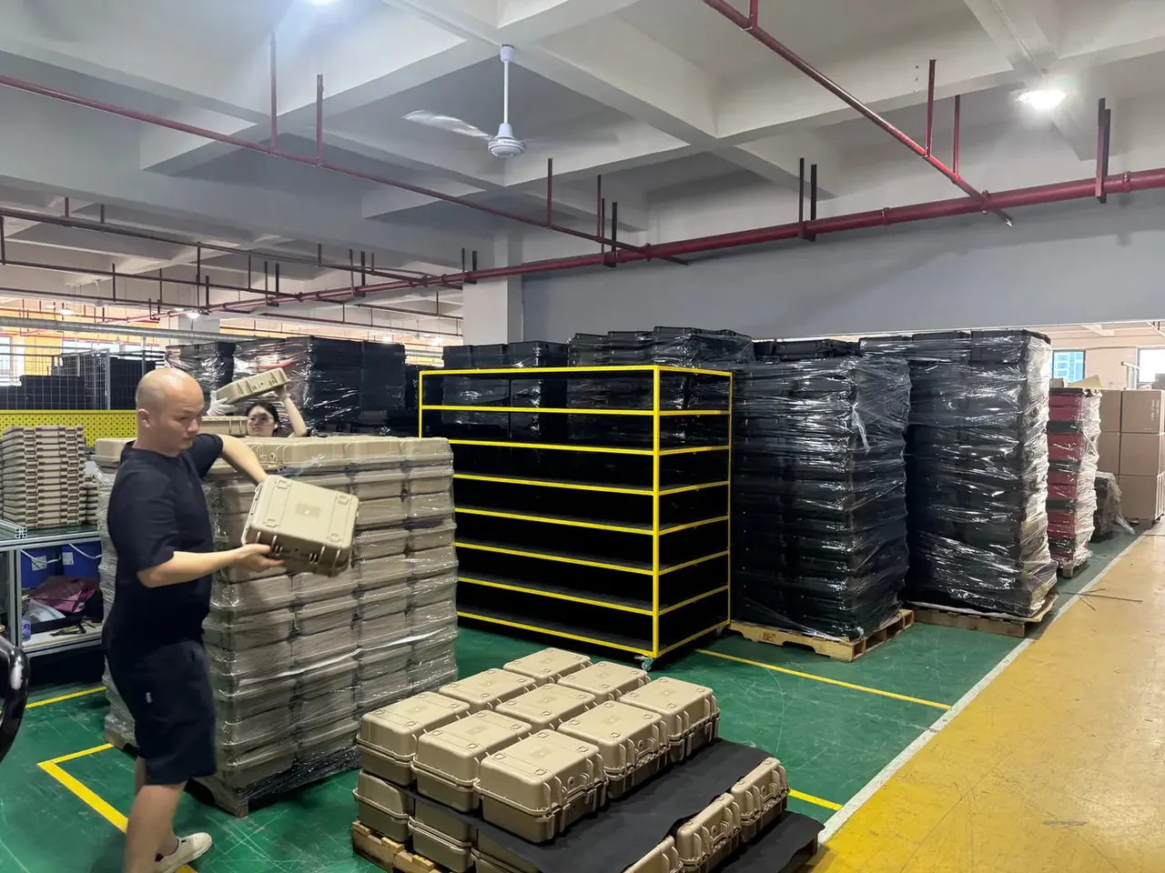 Case Assembly warehouse: a worker in a black T-shirt carrying a tan case, wrapped stacks of tan and black cases, a five-tier yellow-framed rack