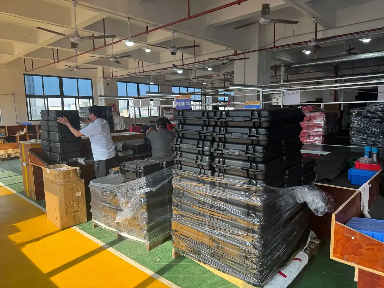 Case Mould: two workers lifting black moulded cases at a timber bench, a blue inspection board above them, film-wrapped stacks and pink crates behind