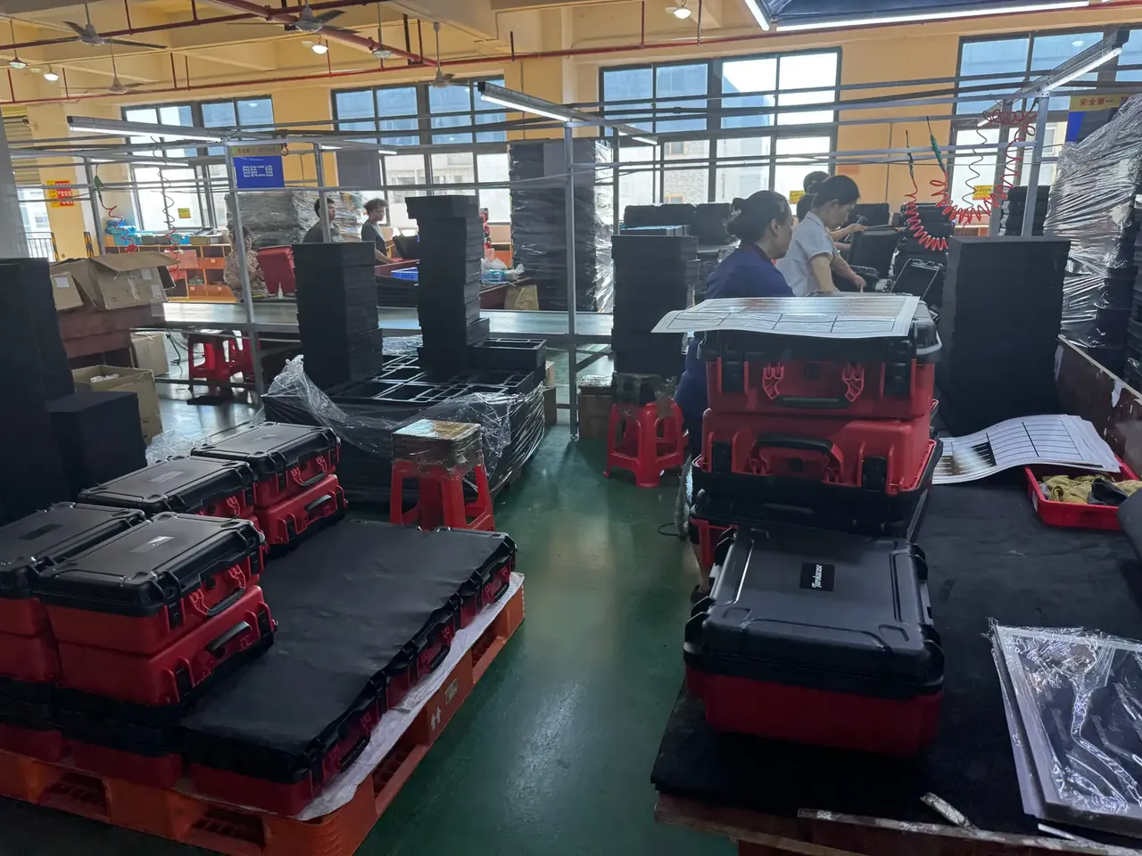 Case Mould: red case bodies stacked with black lids on a pallet, two women working at a bench, tape and label sheets on the table