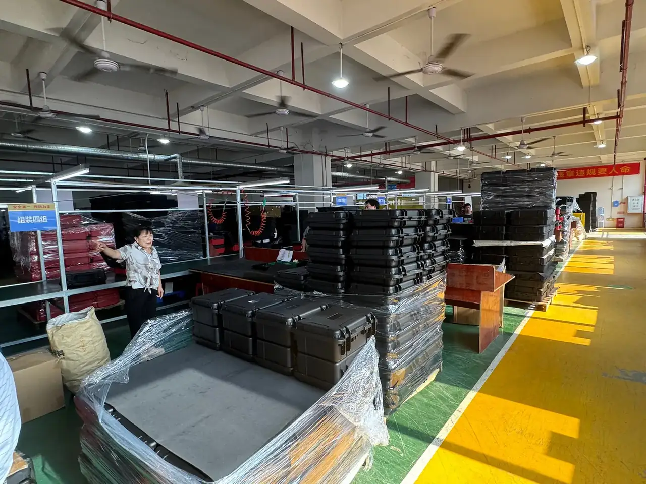 Case Mould: stacks of shrink-wrapped black cases on wooden pallets, green floor meeting a yellow aisle line, a worker raising one hand, red pipe overhead