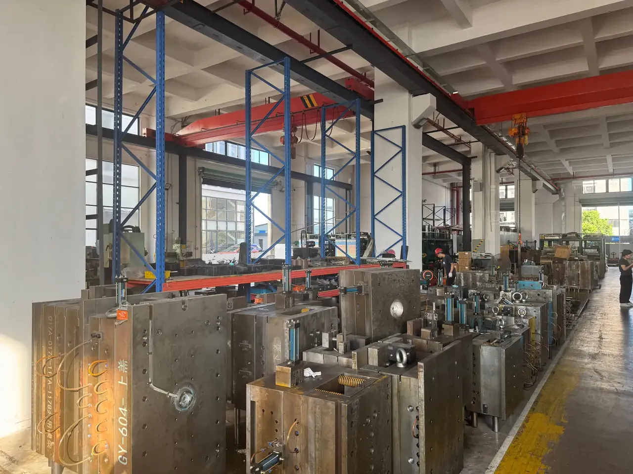箱体模具：同一车间里模具按编号成排摆放，前景钢制模架型腔面朝上，两名工人在通道右侧低头查看，蓝色货架与红色吊车梁在上方
