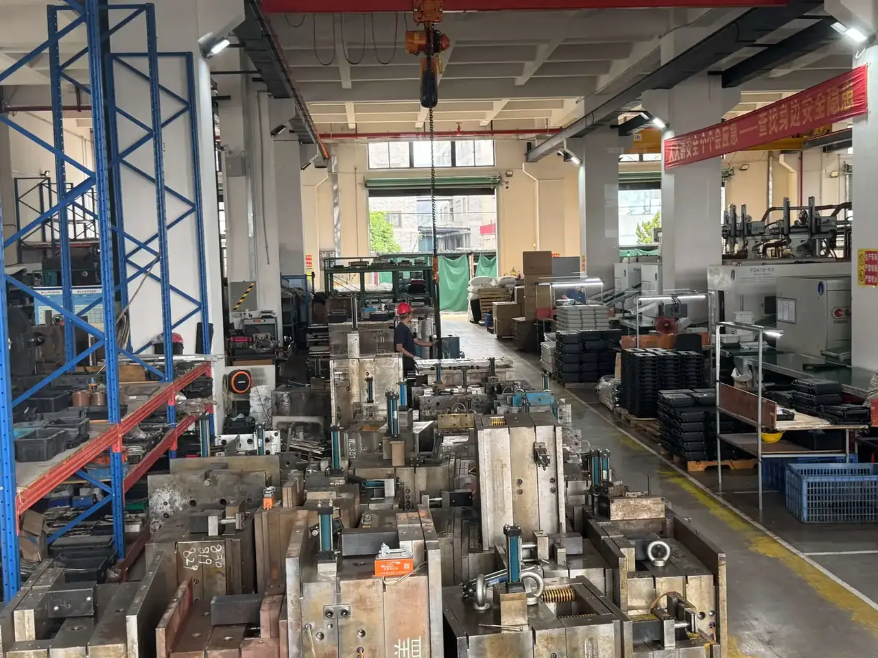 箱体模具：车间通道上方挂着橙色电动葫芦，穿红衣戴红帽的工人站在模具之间，右侧托盘上码着黑色塑料箱，黄色地面线与红色横幅在后方