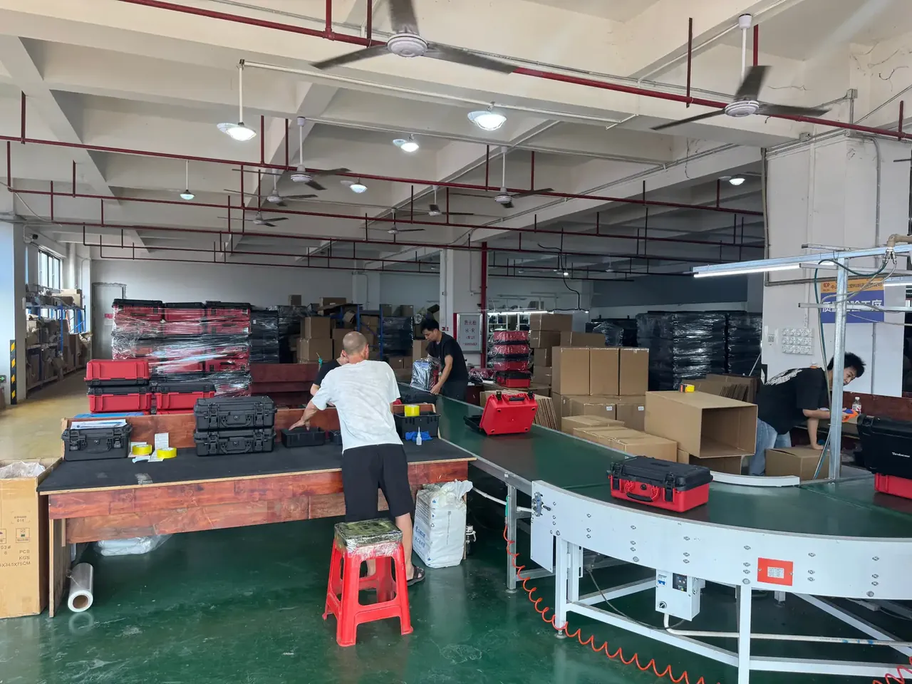 Case Mould: shrink-wrapped black cases on a wooden pallet, green floor by a yellow aisle line, a worker bent over a bench, red pipes above