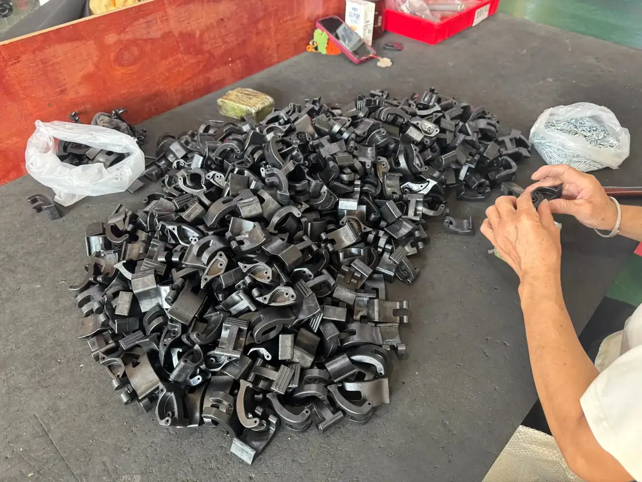 Case Mould: hundreds of black plastic latch parts piled on a grey bench, two hands lifting one part to check flash, white bags beside