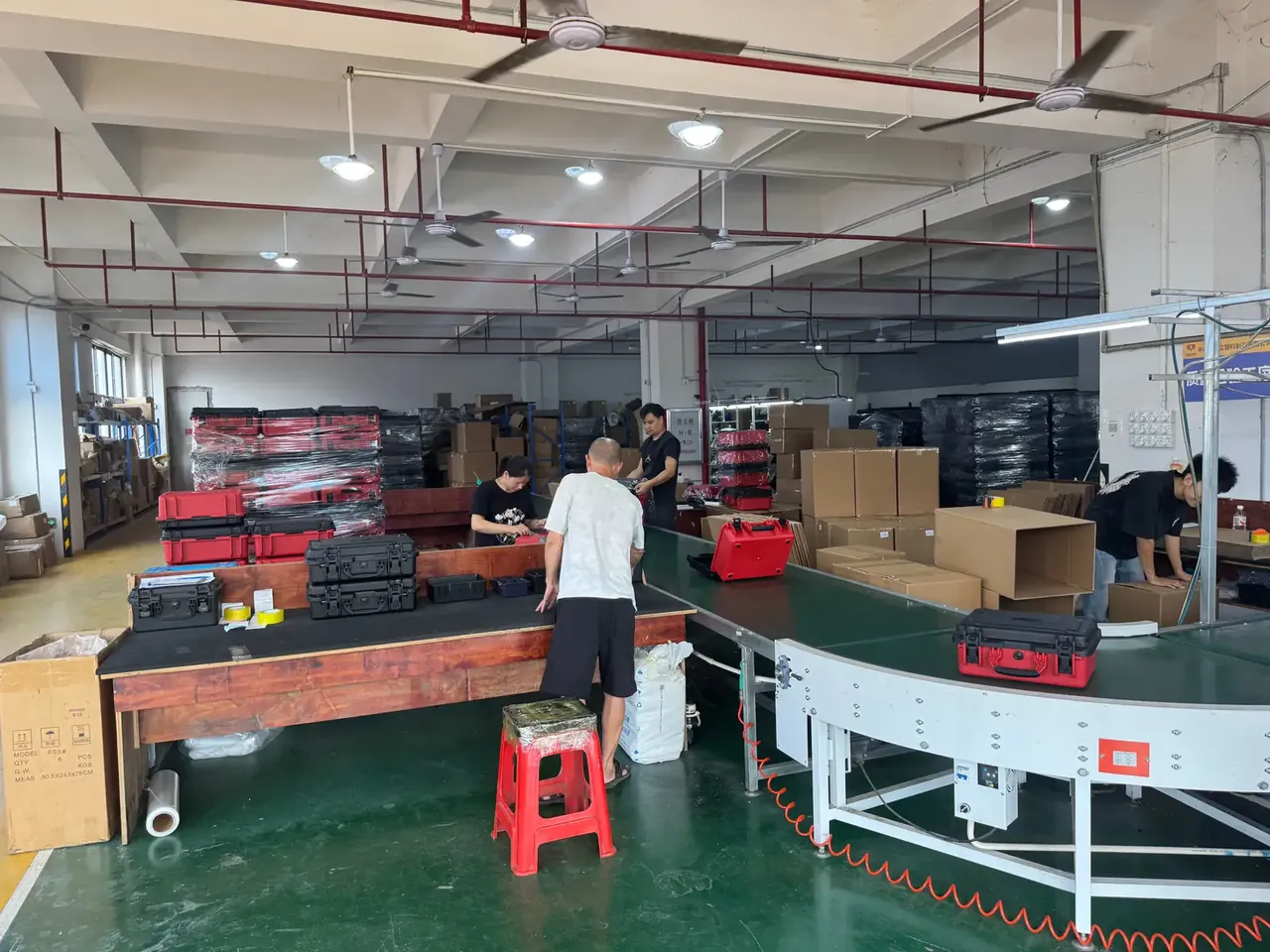 Case Mould: red and black tool cases on wooden benches, a green conveyor curving past, a worker in white beside a red stool