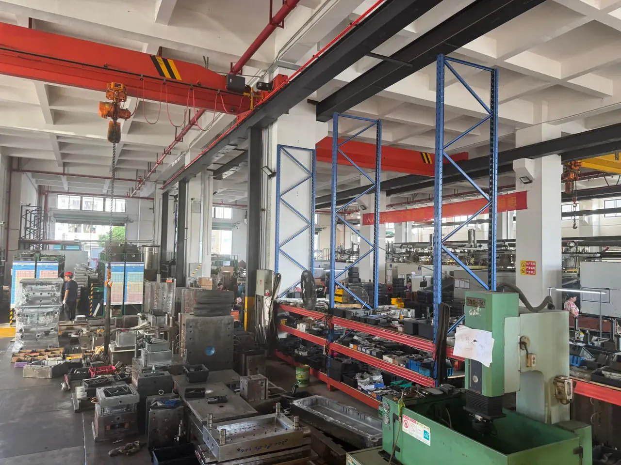 Communication Equipment Case: blue racking mid-shop, a red gantry hook overhead, steel moulds flat on the floor, a green vertical machine at right