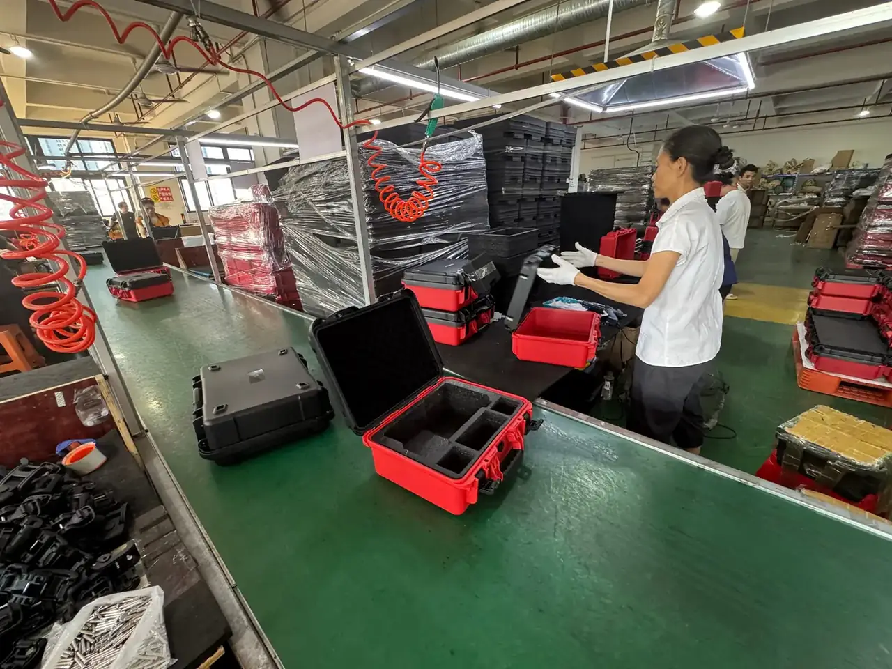 Communication Equipment Case: three open red and black cases on a green bench with black foam dividers, a gloved worker, wrapped pallets behind