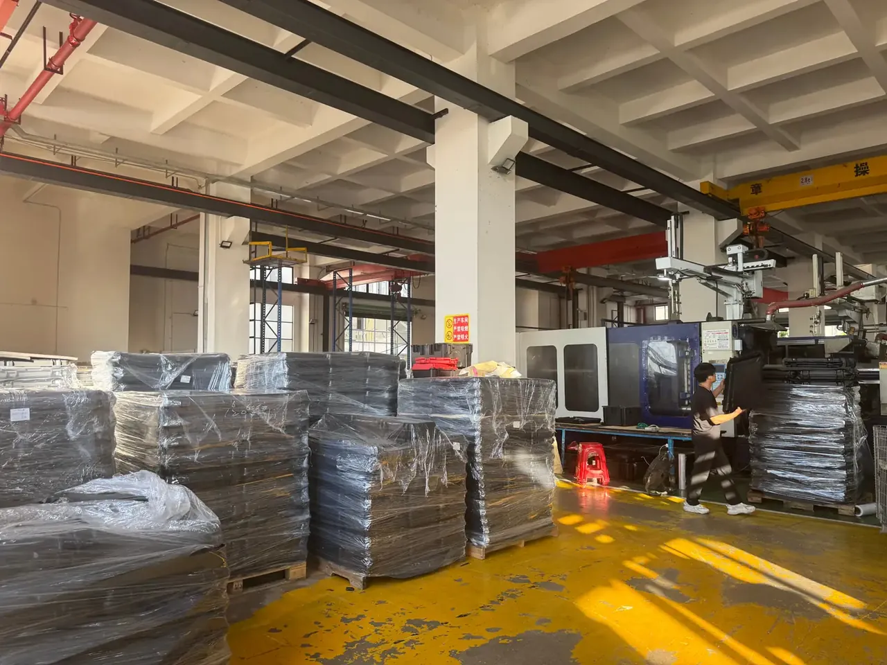Custom Protective Case Manufacturer: yellow workshop floor, pallets of dark cases under clear film, a worker carrying a case toward a blue press