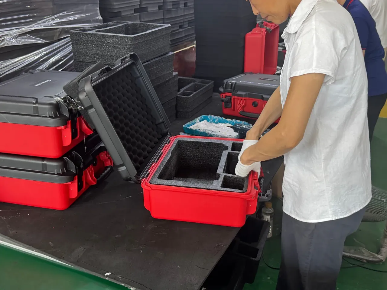 Custom Protective Case Quotation: gloved worker pressing grey foam into a red case on a bench, red and black cases stacked left, wrapped pallets behind