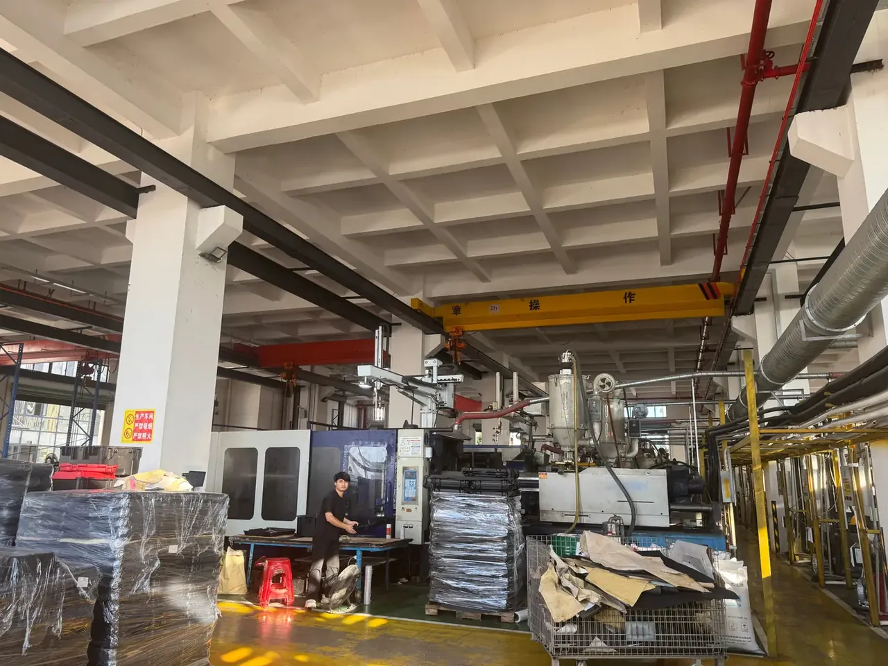 Injection Molded Plastic Box Manufacturer: worker in black beside a large grey injection press, yellow crane girder overhead, film wrapped pallets and a red stool