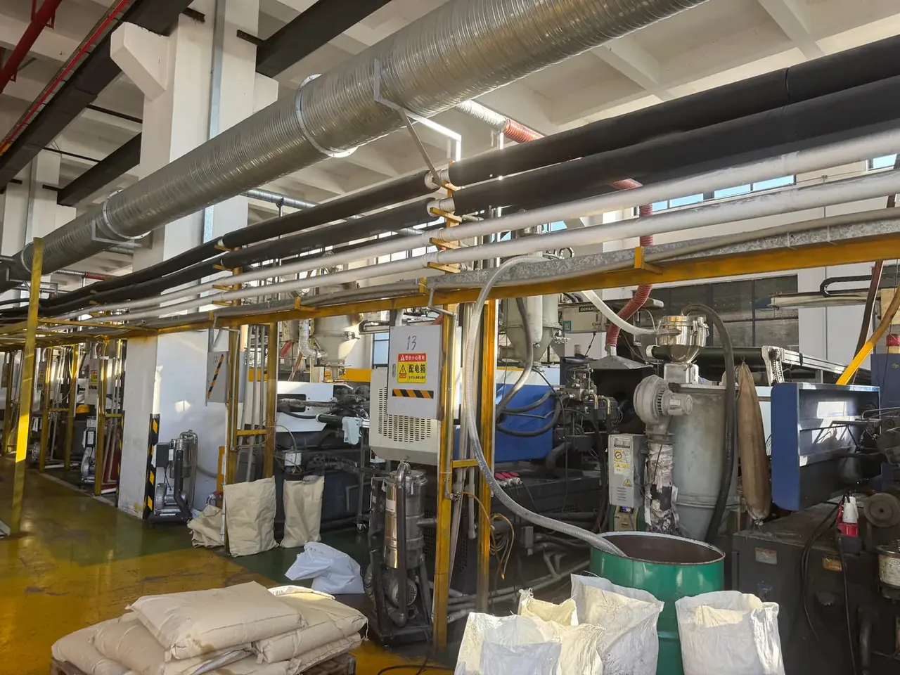 Injection Molded Protective Case: blue and white injection machines behind yellow guards, corrugated ducts overhead, white resin sacks and green drums on the floor