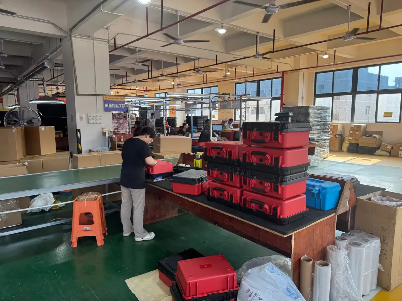 Injection Molded Protective Case: a worker opening a red case on a green conveyor, four red and black cases stacked on a bench behind