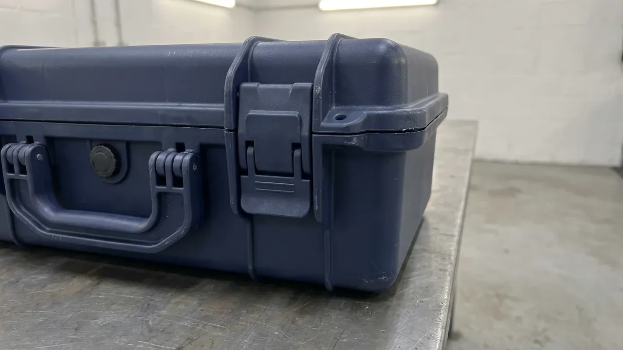 Moisture-Resistant Equipment Storage Case: navy case closed on a metal bench, round vent cap, two latches and folded handle on the front, factory wall behind