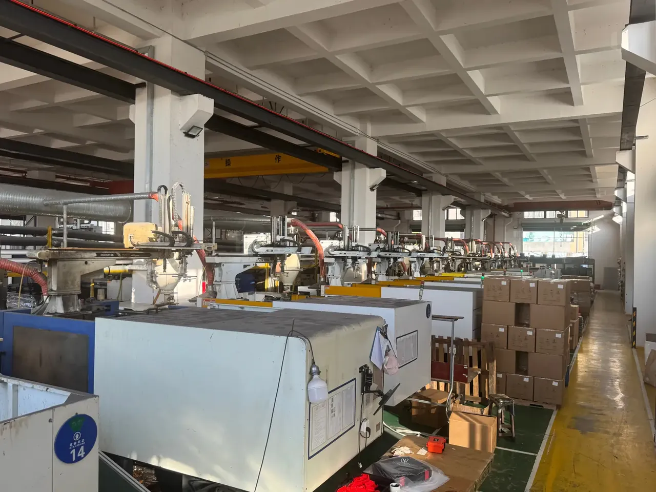 ODM Protective Case: a row of horizontal injection moulding machines receding into a workshop, hoppers fed by red hoses, a crane beam overhead, stacked cartons on the right, yellow floor lines