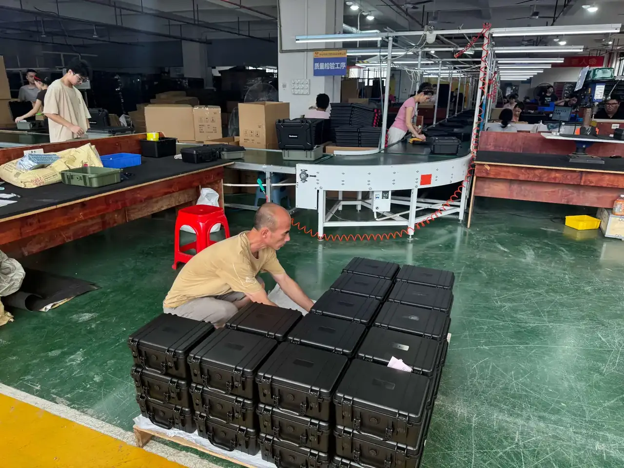 Pistol Case: black cases stacked four high on a green floor, a worker in a beige shirt crouching to align lids, conveyor behind