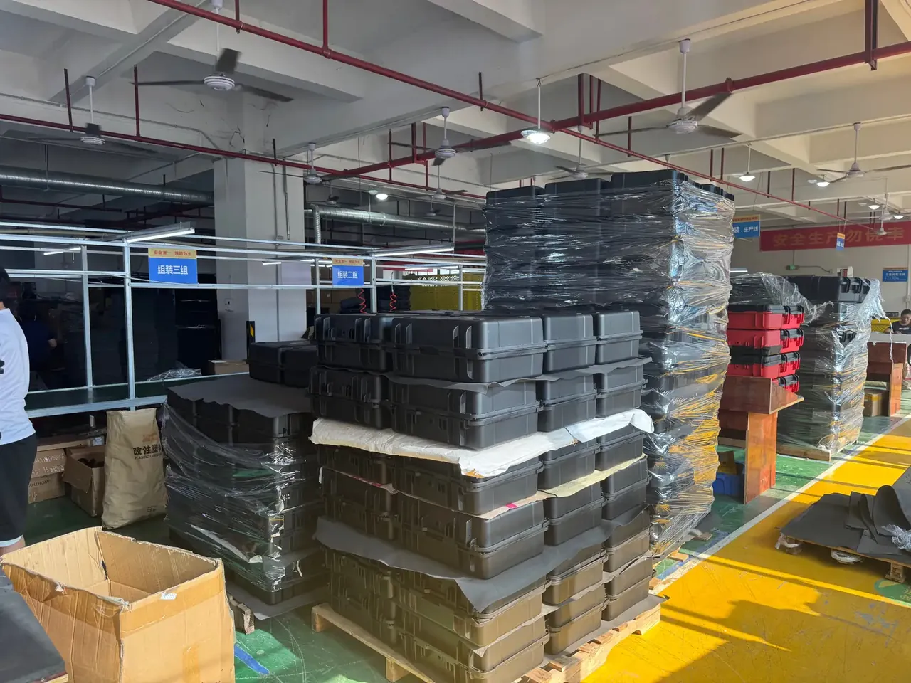 Plastic box factory: black cases stacked on wrapped pallets, a red stack beside them, blue station signs on a yellow floor line
