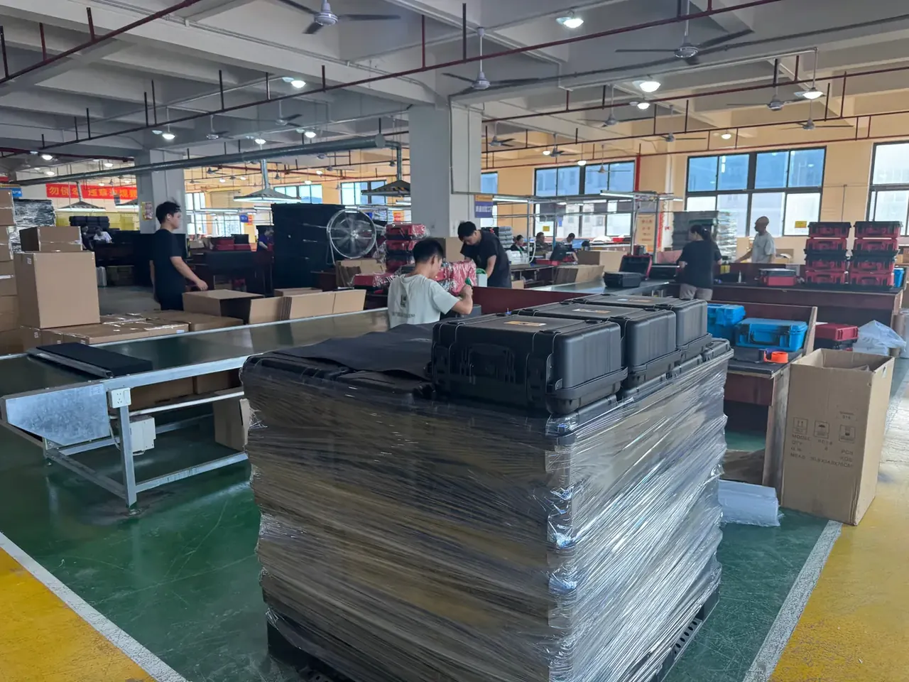 塑料箱批发供应商：包装车间绿色地坪上，一垛纸箱被透明缠绕膜裹了多层，垛顶并排放着三只黑色手提箱，后方输送带旁有几名工人在打包，右侧立着印字的牛皮纸箱