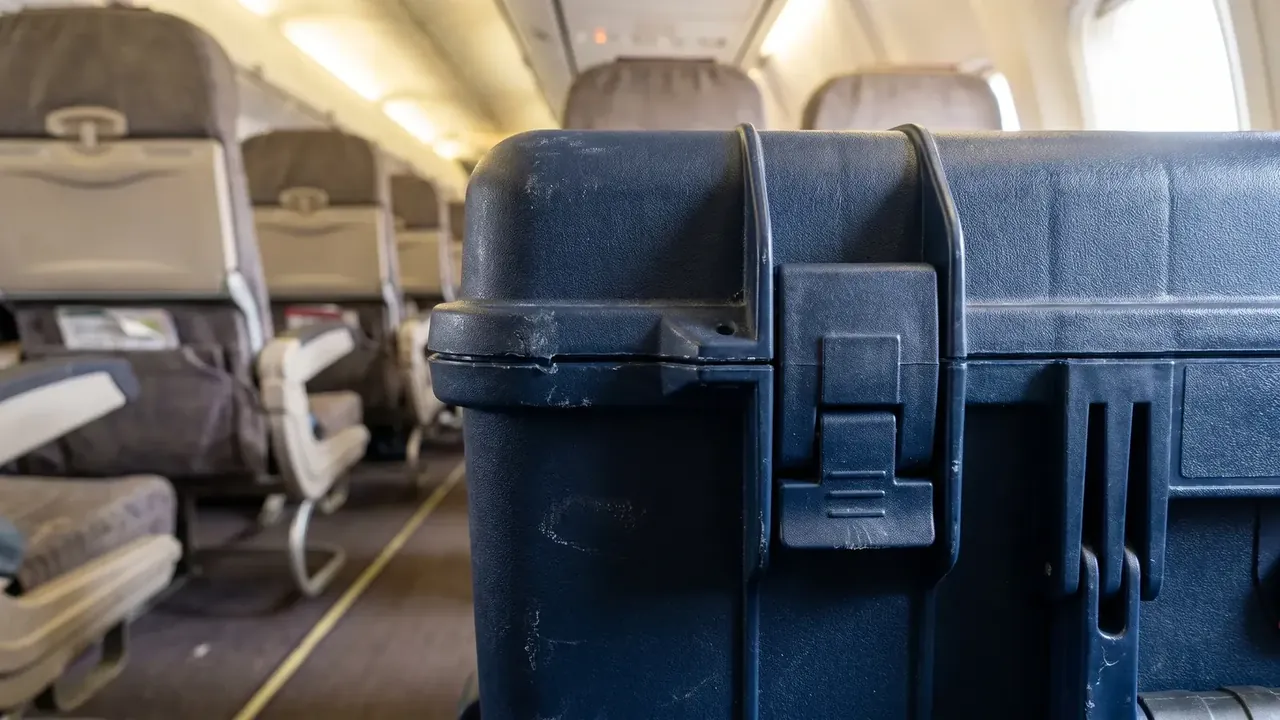 Plastic suitcase with wheels wholesale navy case standing in an aircraft aisle, latch and corner close to the lens, seat rows and overhead bins behind