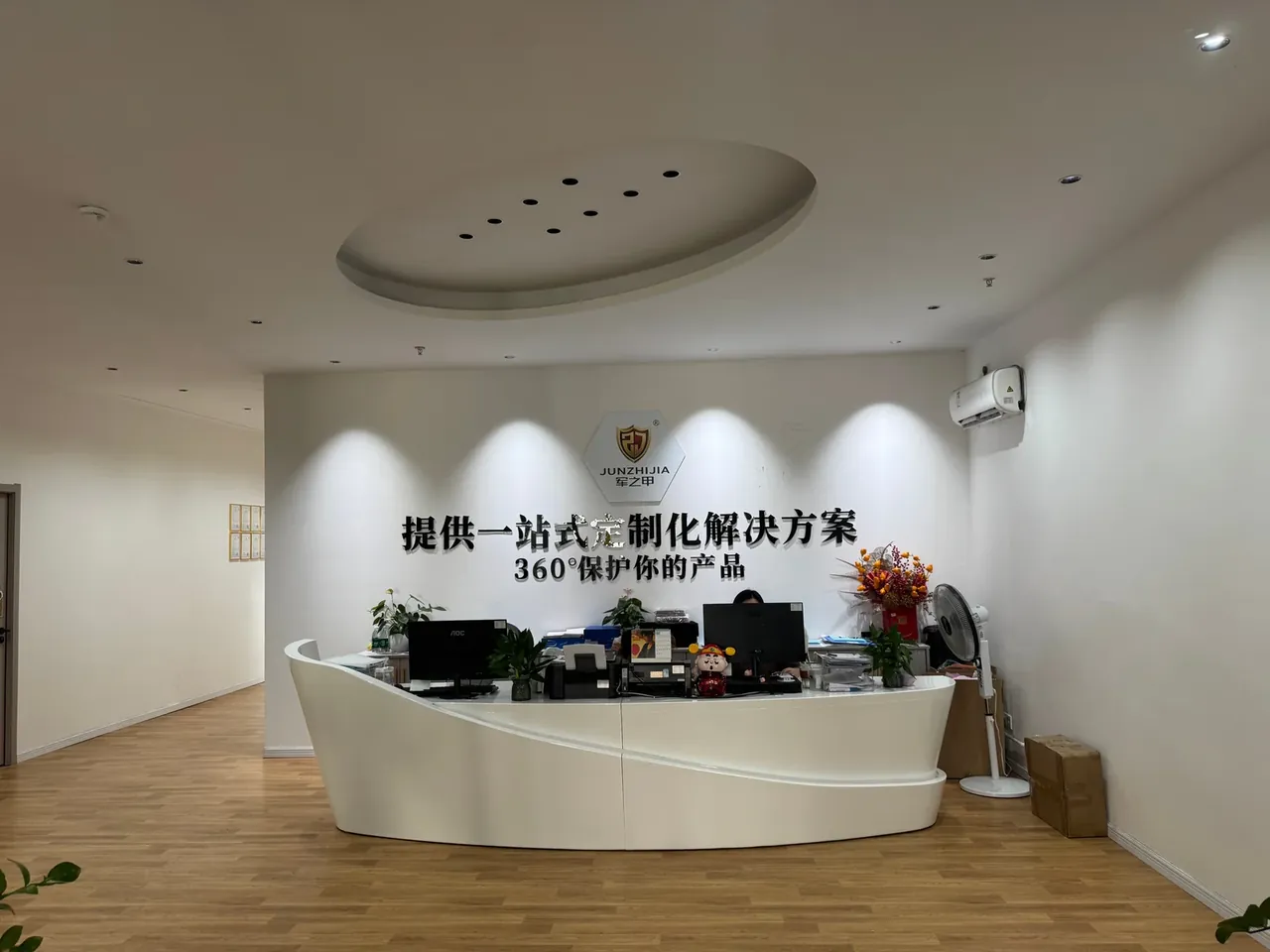 Plastic Tool Box, Portable: a curved white reception desk below a shield-shaped logo wall and a round ceiling cove, with plants and a fan