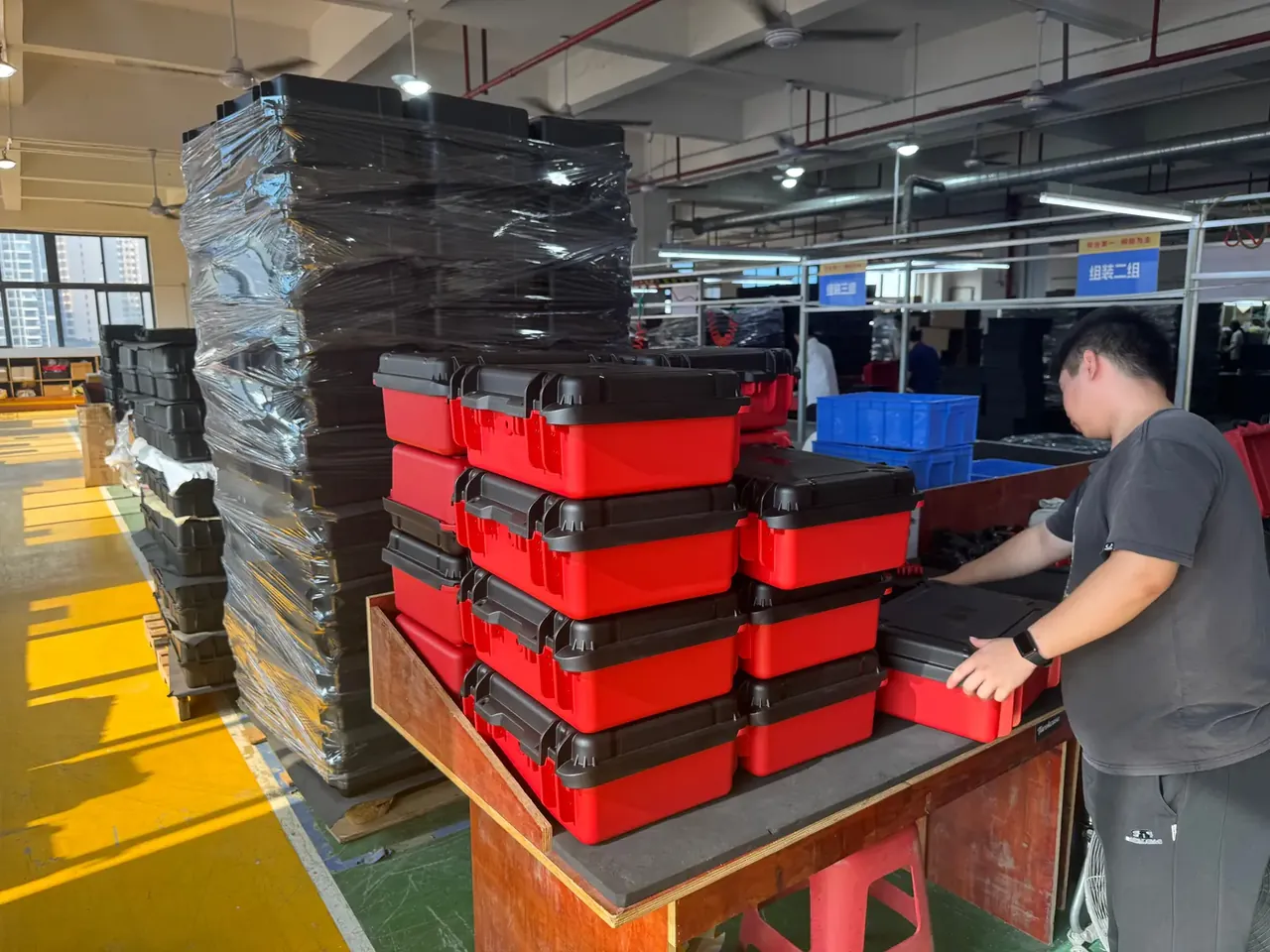 Portable Tool Box: red bodies with black lids stacked six high on a bench, a worker steadying the top case, wrapped stacks behind