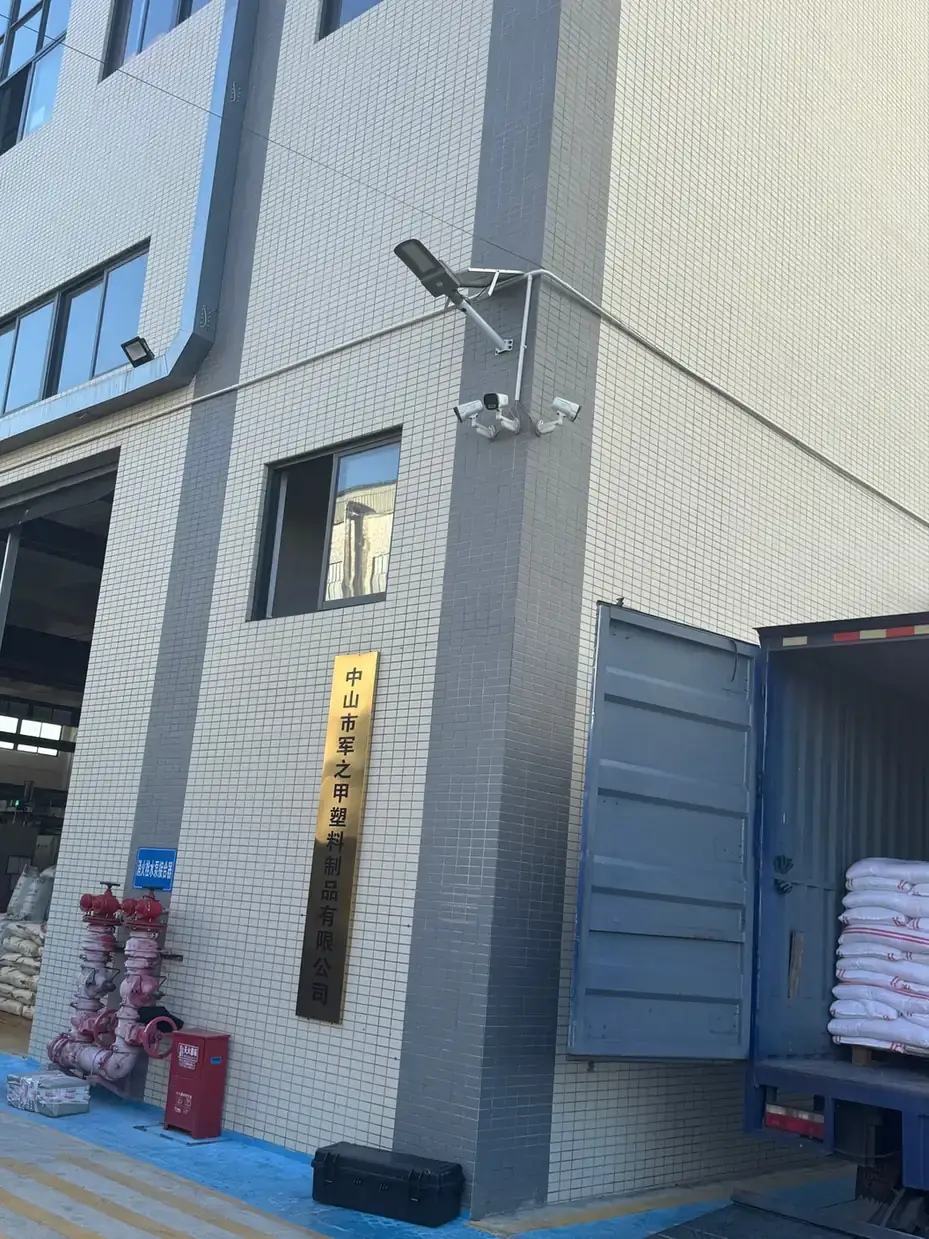 Protective box: black long box lying on blue-painted ground beside a freight truck, pale tiled factory wall with a gold sign and two cameras