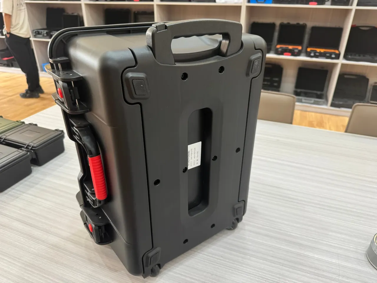 Protective box: black box standing upright on a table showing its back, folding top handle, telescopic red-grip handle at left, two wheels, sample shelves behind