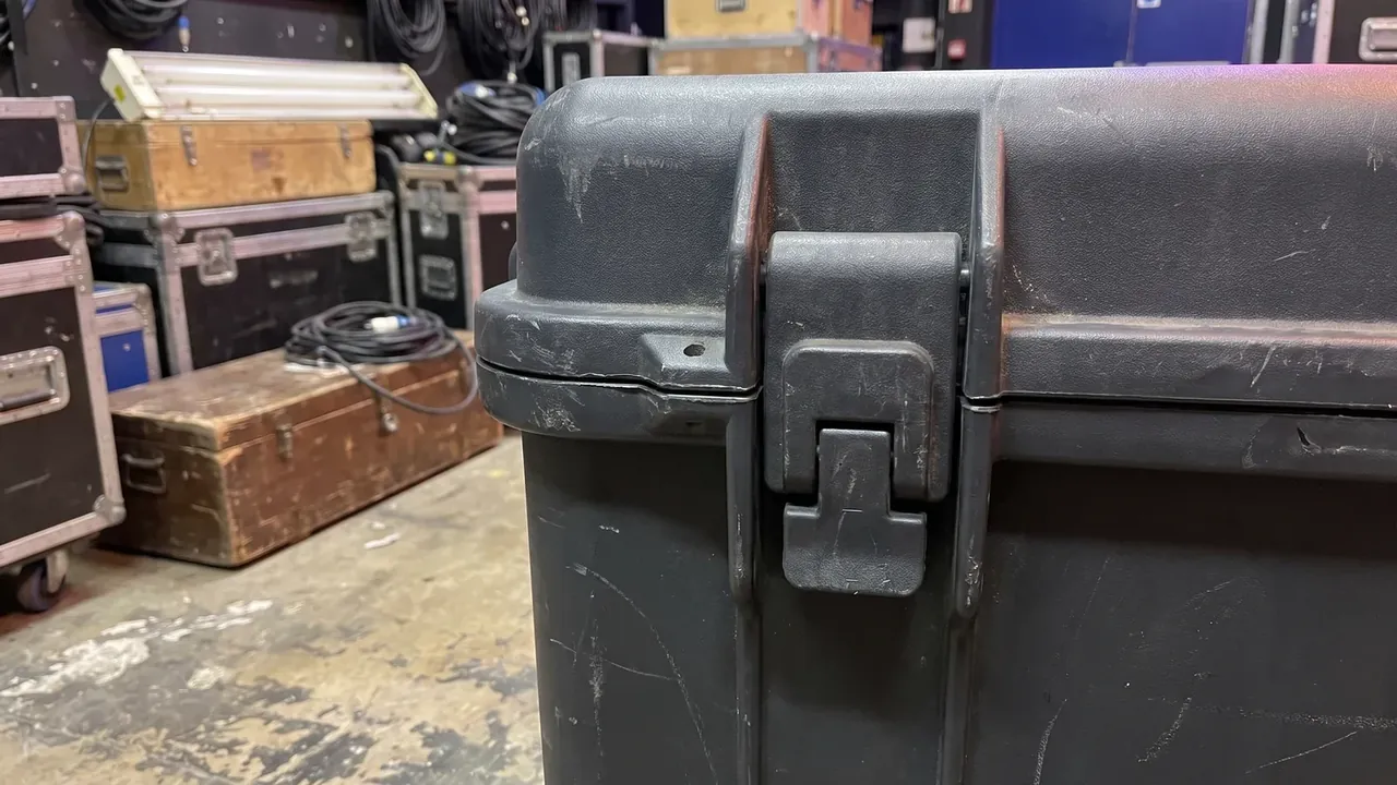 Road Case corner close-up, the latch tongue half open, fine scratches across the shell, backstage cases and floor out of focus