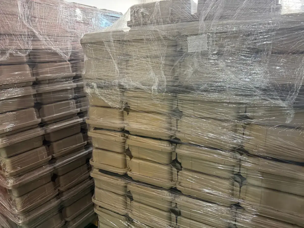 Rotomolded Protective Case Manufacturer: warehouse stacks of khaki cases nested eight high, wrapped in clear stretch film, a darker stack at left on a concrete floor