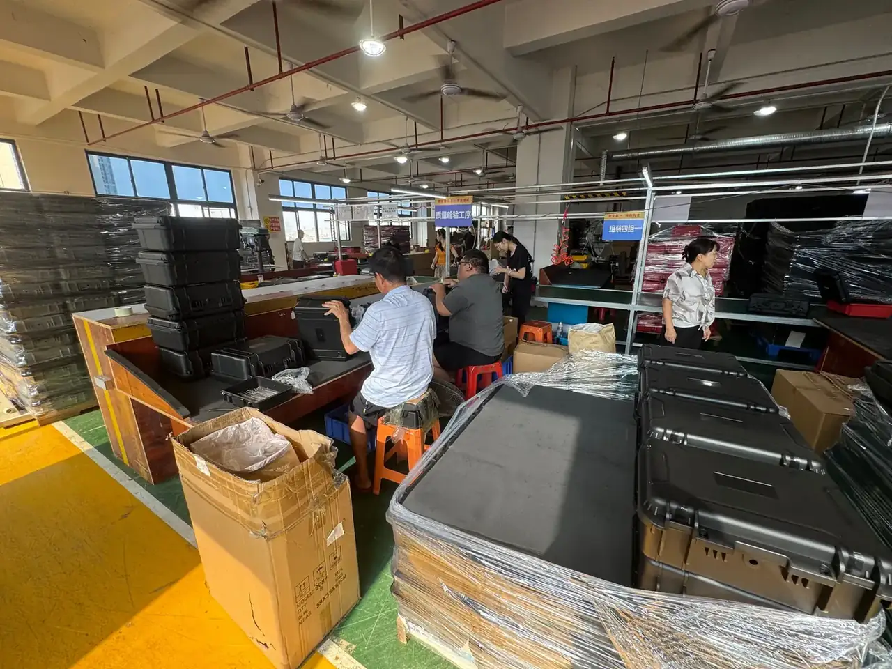Rotomolded Protective Case Manufacturer: workers on orange stools finishing black cases at benches, a stack of black cases at left, two blue overhead signs, yellow floor markings