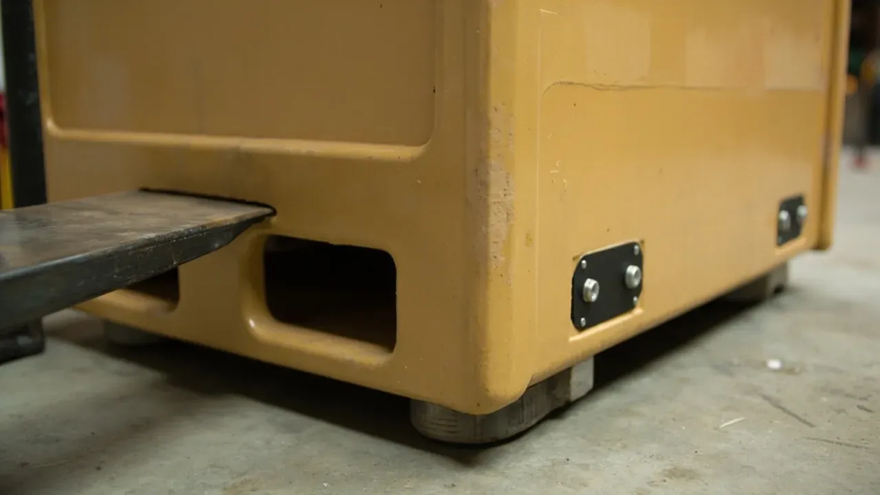 A forklift fork entering the moulded pocket in the case base, a bolted lifting plate and castors below