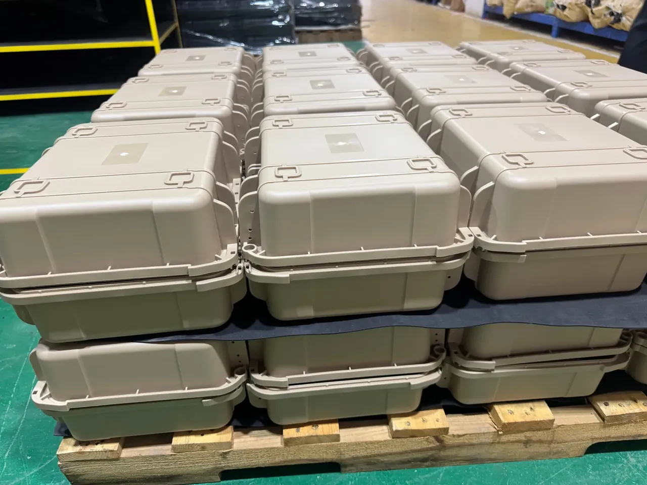 Tool Bag: beige hard cases in rows on a wooden pallet, black separators between layers, green factory floor behind