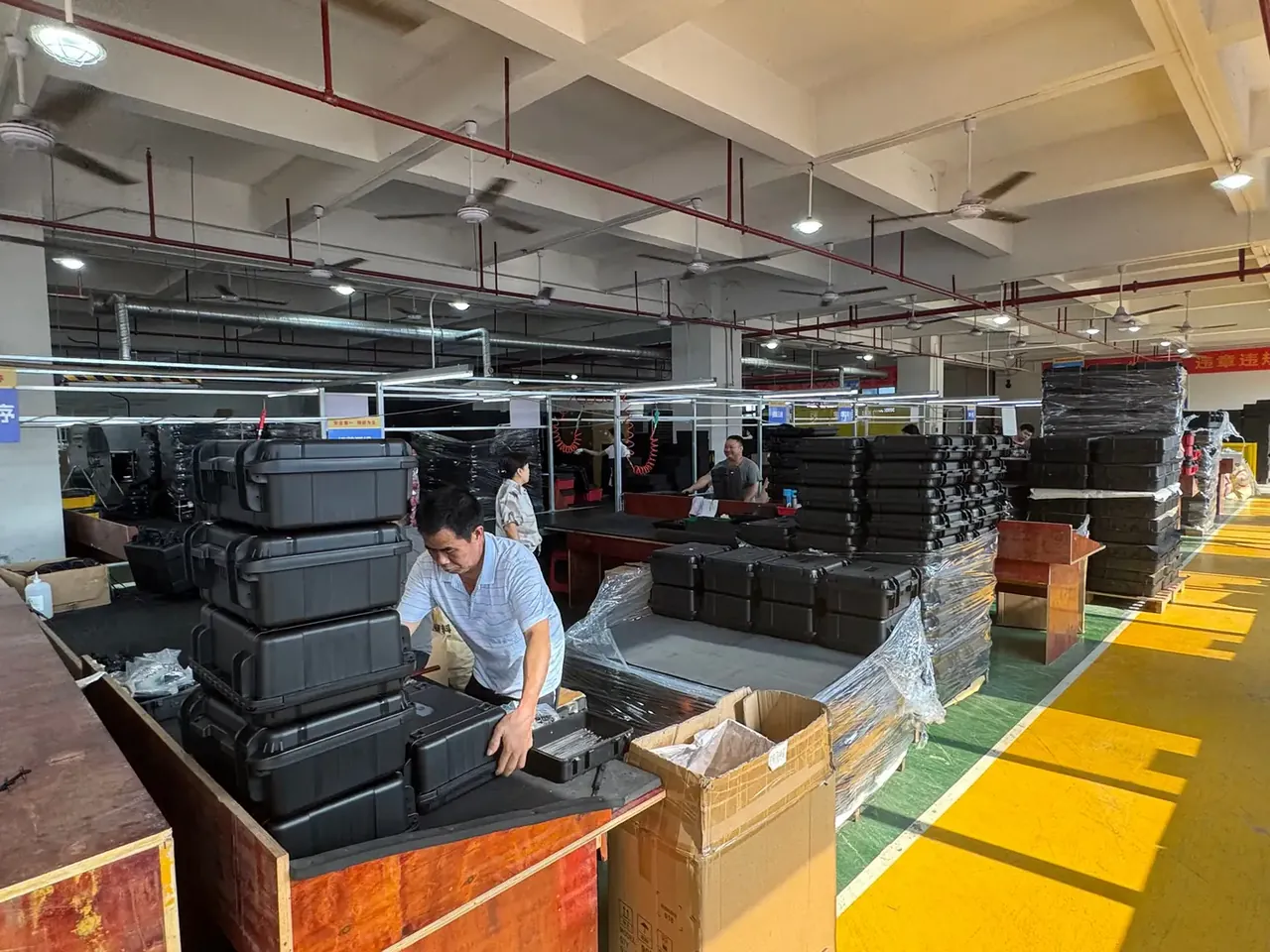 Tool Box: yellow floor lines in a packing hall, black rotomoulded cases stacked on timber pallets, two workers handling them, wrapped pallets right
