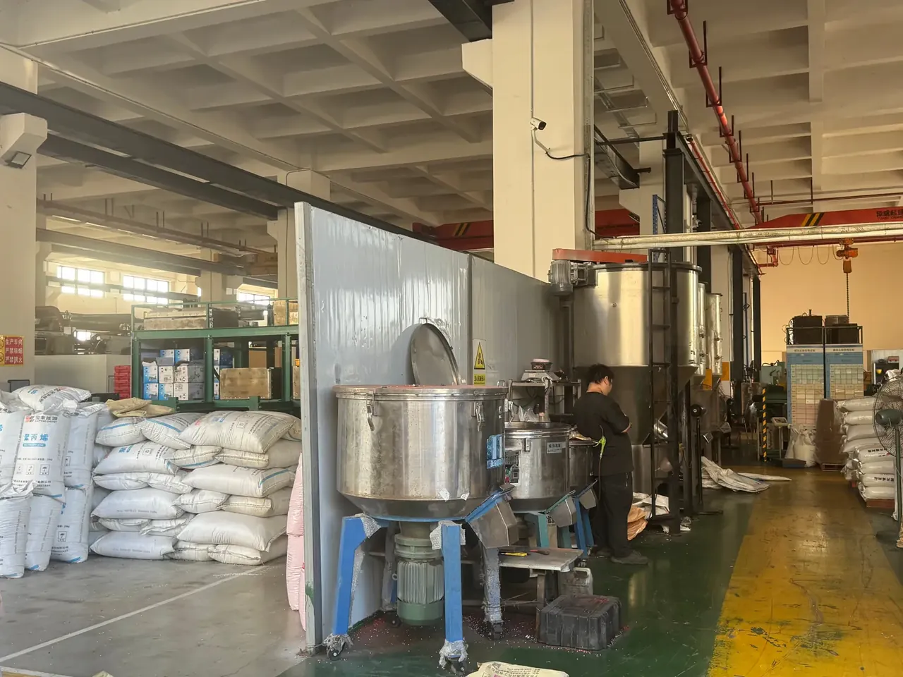 Tool Box Wholesale: stacked white resin sacks beside open stainless mixing tanks, a worker in black standing by, green floor with yellow lines