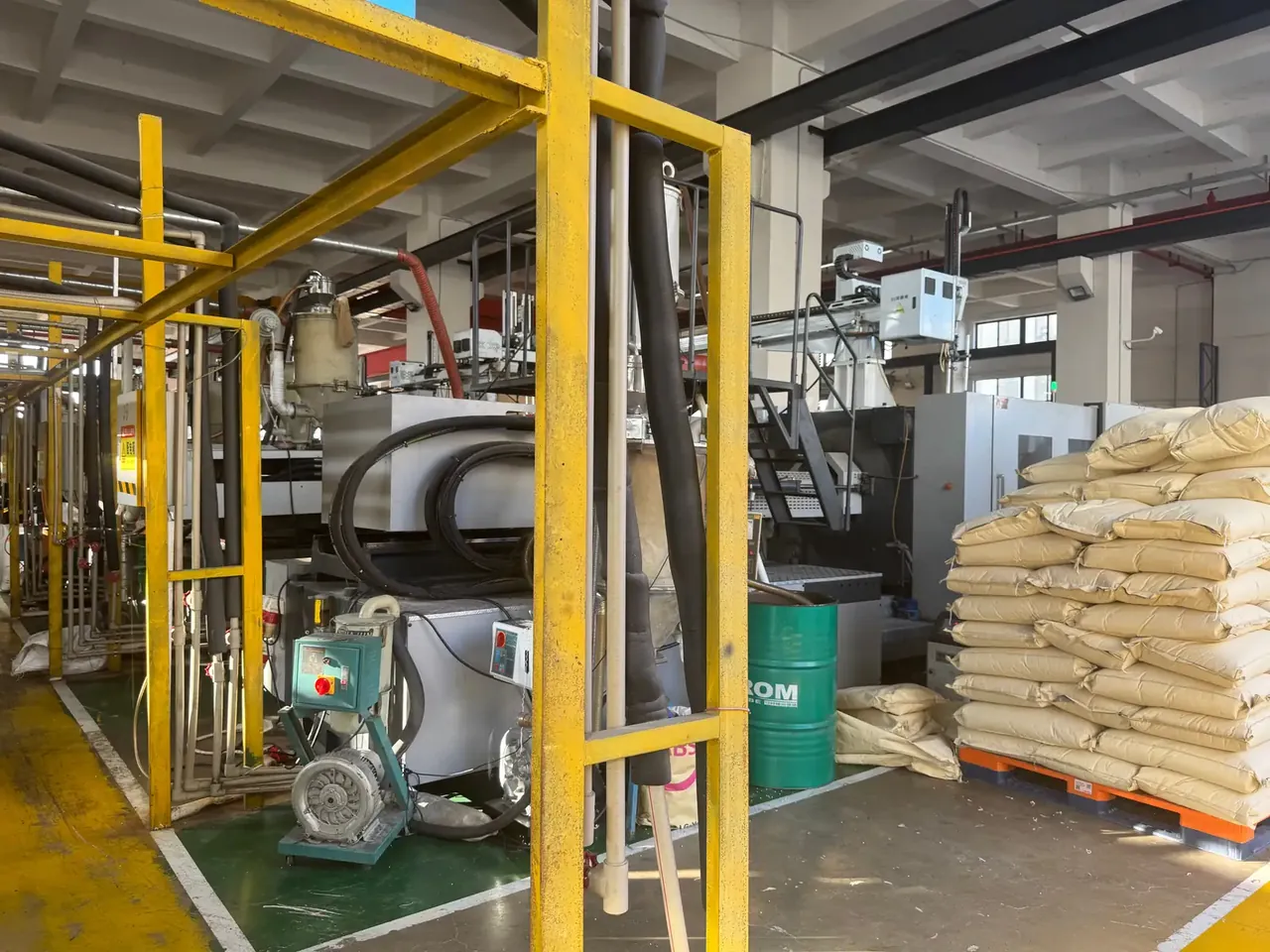 Tool Box Wholesale: yellow steel framing in front of an injection machine, black hoses, a green drum beside stacked beige resin bags on a pallet