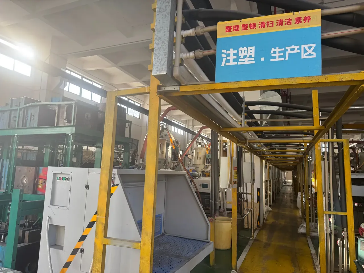 工具携带器：注塑车间入口上方挂着蓝底白字生产区标牌，黄色钢管框架下排列着绿色注塑机与模具，地面画着黄色通道线