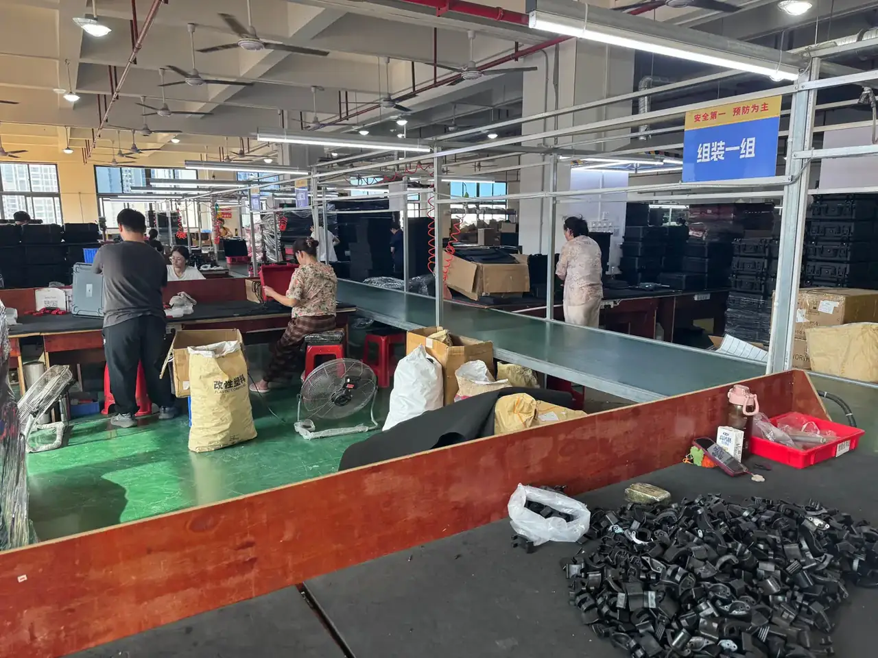 Tool Carrier: blue assembly sign over a green-floored bench row, four workers at long tables, sacks stacked beside