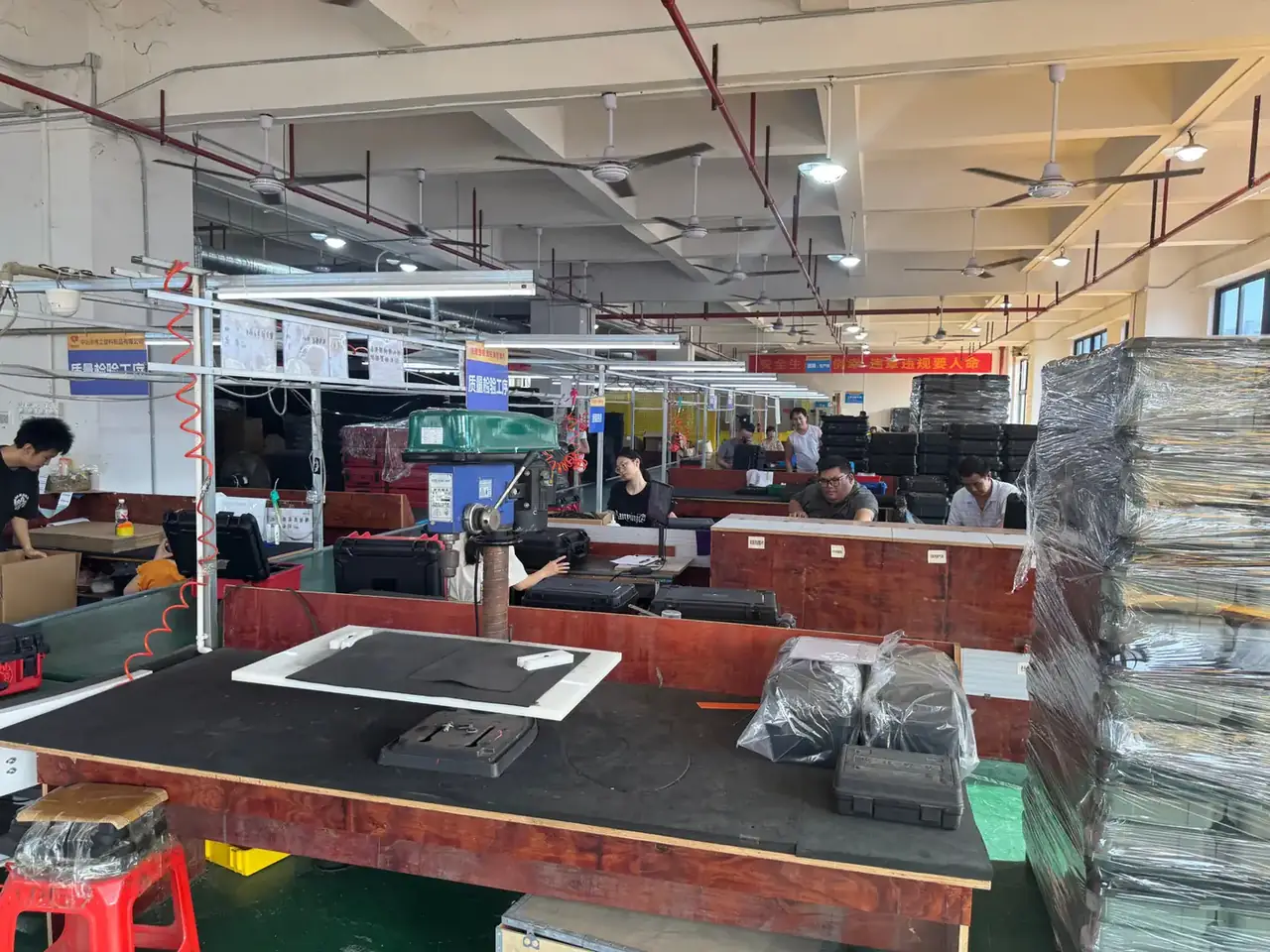 Tool Chest: workshop benches with black cases, workers in black shirts seated, wrapped pallets stacked at right, ceiling fans and tube lights