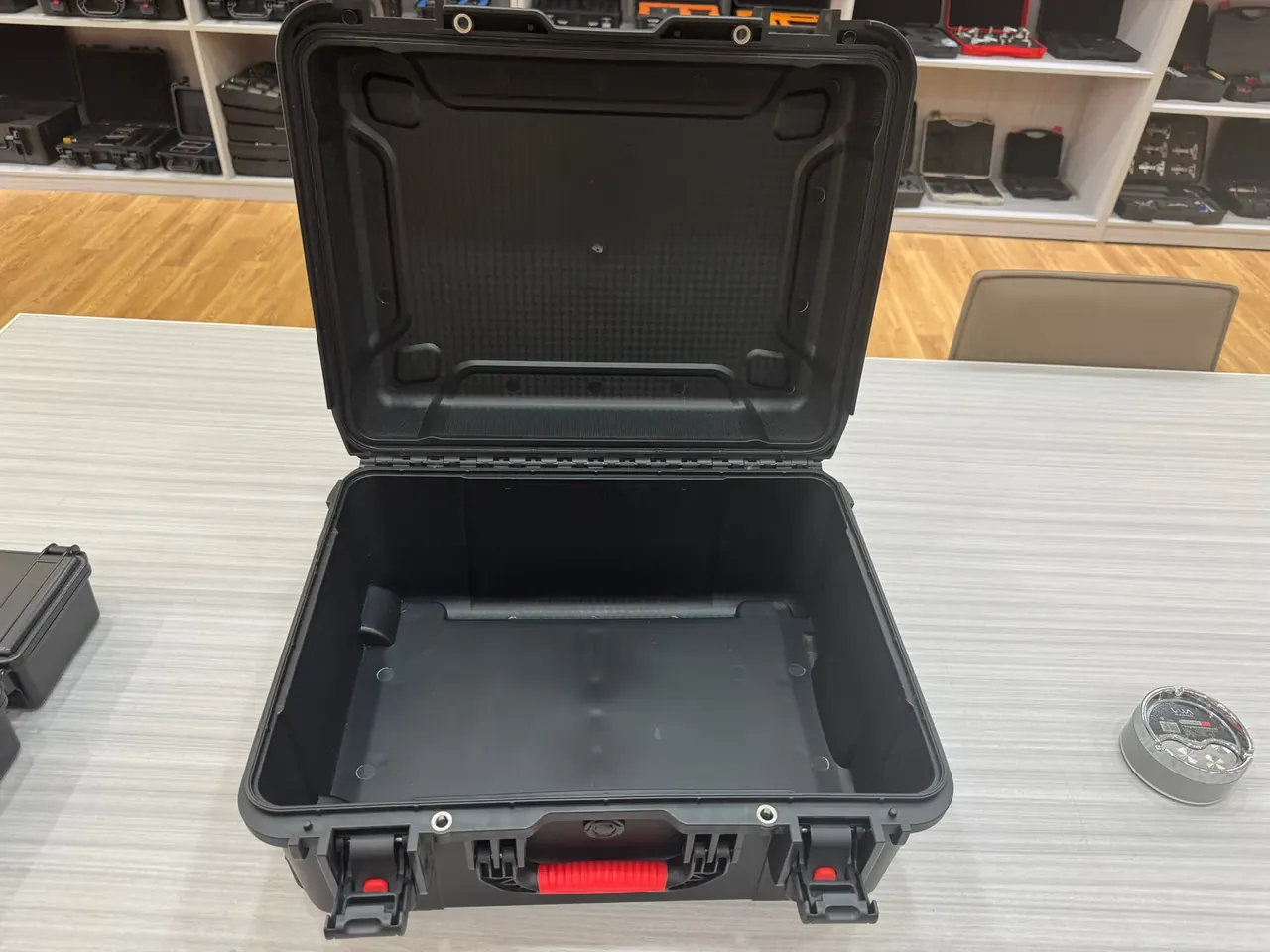 Tool Kit Chest: black case open on a wood-grain table, red latch and two silver twist locks, shelves of cases behind, a tape measure