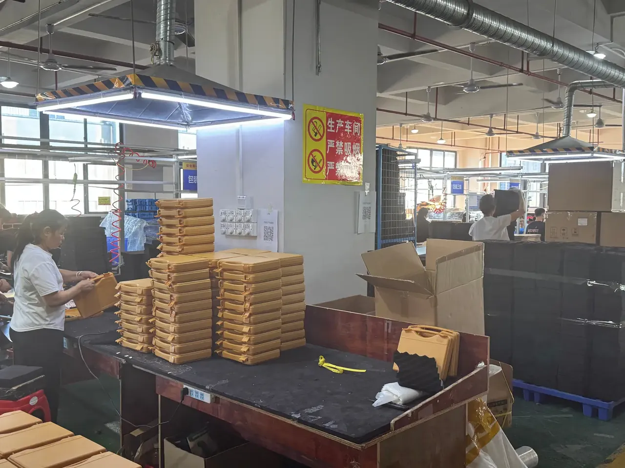 工具卷：车间包装工位上堆着几摞牛皮纸色纸盒，一名女工站在黑色台面前整理盒盖，右侧是敞口大纸箱与缠膜的黑箱堆，柱上挂着黄色禁烟牌