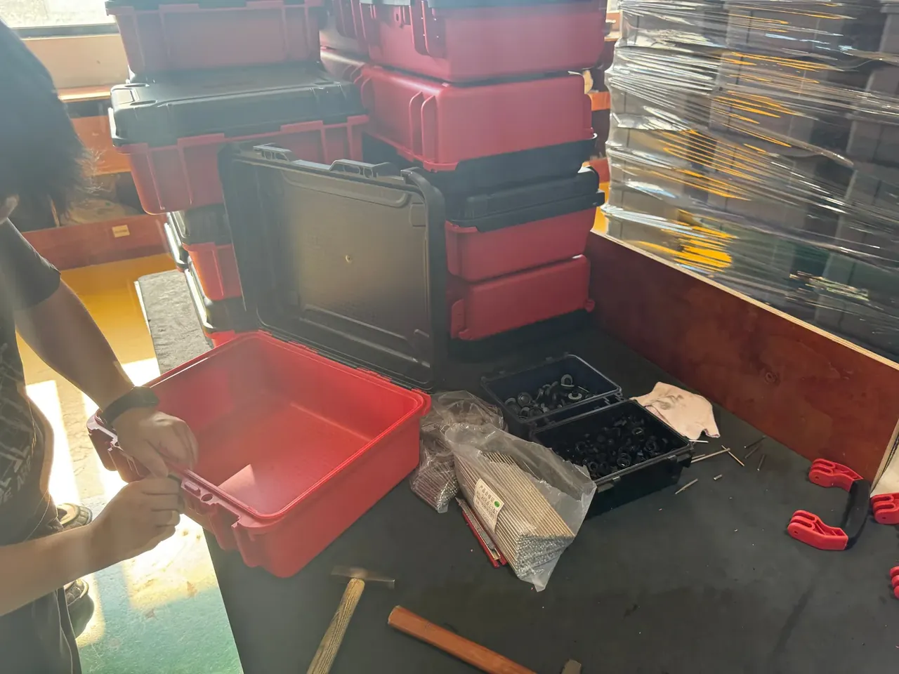 工具卷：工人正掀开一只红色箱盖，桌面上摆着两格黑色零件盒、成袋的五金配件与一把锤子，身后红黑两色箱体叠到齐腰高