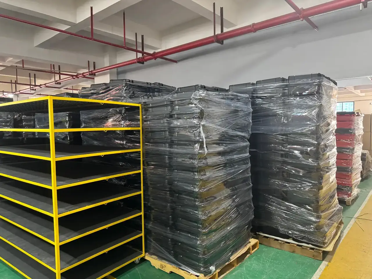 工具卷：仓库里立着一组黄色金属货架，旁边木托盘上码着立柱般的黑色箱体，整垛缠着透明缠绕膜，地面刷成绿色