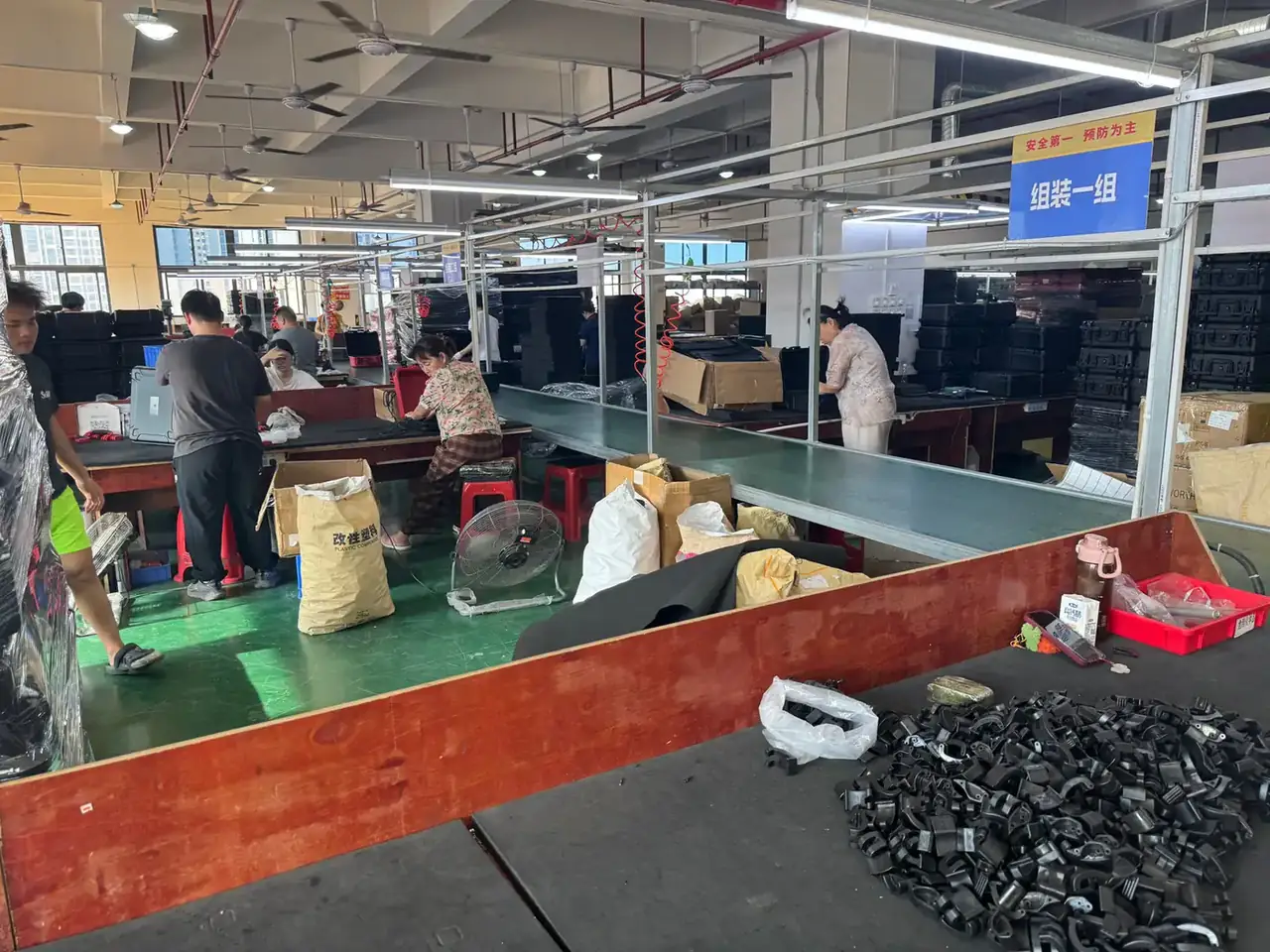 Tool Roll: a steel-framed assembly bay with a blue sign reading assembly group one, workers along green benches, black plastic catches heaped in front