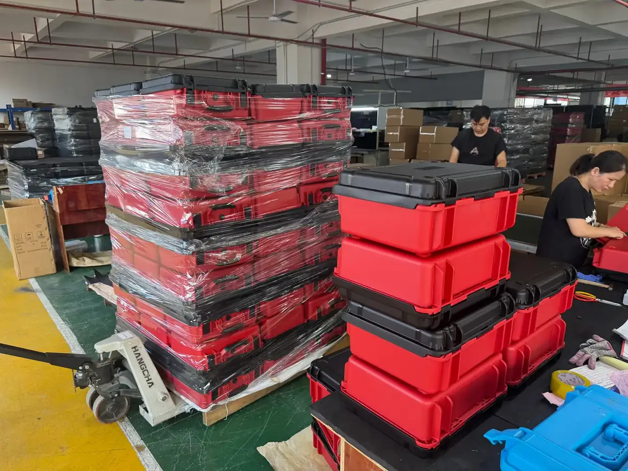 Waterproof case: stacks of red cases wrapped in clear film on pallets, workers sorting at a bench, a pallet truck at left